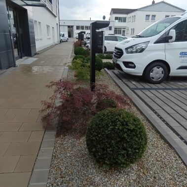 Eingangsbereich eines modernen Firmengebäudes mit gepflastertem Gehweg und bepflanztem Kiesbeet. Darin stehen kugelförmig geschnittene Sträucher und ein roter Zierahorn. Daneben sind Parkplätze mit Firmenfahrzeugen und eine Ladesäule für Elektroautos zu sehen.