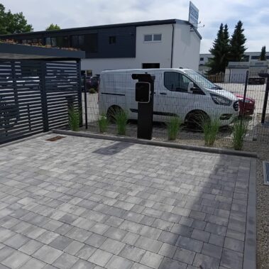 Neu gepflasterter Parkplatz mit grauen Steinen, daneben eine schwarze Ladestation für Elektrofahrzeuge. Im Hintergrund stehen ein weißer Transporter, weitere Autos und ein modernes Firmengebäude.