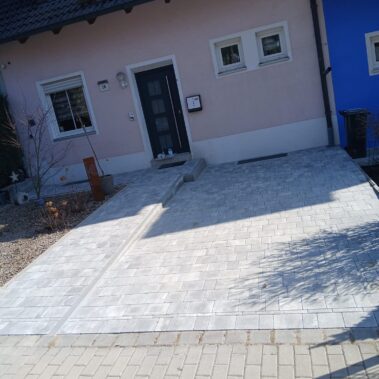 Eingangsbereich des Hauses mit frisch gepflasterter Fläche aus hellgrauen rechteckigen Steinen. Der Bereich wirkt sauber und eben, mit integriertem schmalem Ablaufkanal. Links führt ein kleiner Weg zur Haustür.