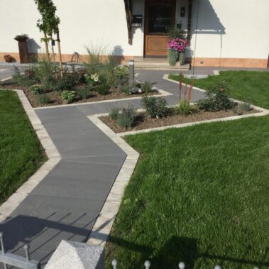 Vorgarten mit frisch angelegtem Rasen und modernem Plattenweg, der zum Hauseingang führt. Beete mit jungen Sträuchern, Gräsern und Blumen sind in geometrischen Formen eingefasst. Vor der Haustür stehen Pflanzkübel mit blühenden Blumen.
