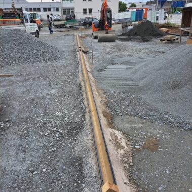 Baustelle mit Schotterfläche, auf der ein Bagger und Baumaterialien stehen. Im Vordergrund verläuft eine vorbereitete Entwässerungsrinne aus Metall.