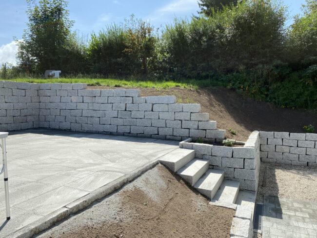 Neu angelegte Terrasse mit hellen Steinplatten und angrenzender Stützmauer aus grauen Blocksteinen. Eine Treppe führt von der Terrasse nach unten, daneben ist ein Bereich mit Kies und Pflanzsteinen.