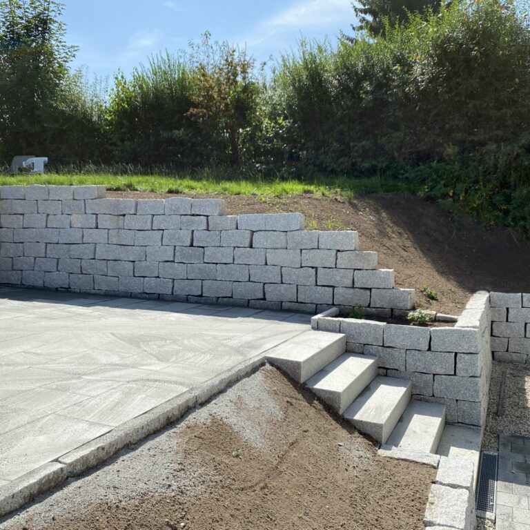 Neu angelegte Terrasse mit hellen Steinplatten und angrenzender Stützmauer aus grauen Blocksteinen. Eine Treppe führt von der Terrasse nach unten, daneben ist ein Bereich mit Kies und Pflanzsteinen.