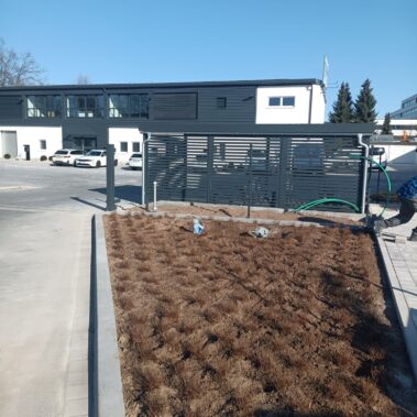 Neu angelegtes Firmengelände mit gepflasterter Zufahrt und einem großen Beet am Rand. Das Beet ist frisch bepflanzt mit kleinen Sträuchern in gleichmäßigem Raster. Im Hintergrund stehen Firmengebäude, Autos und eine Sichtschutzwand.