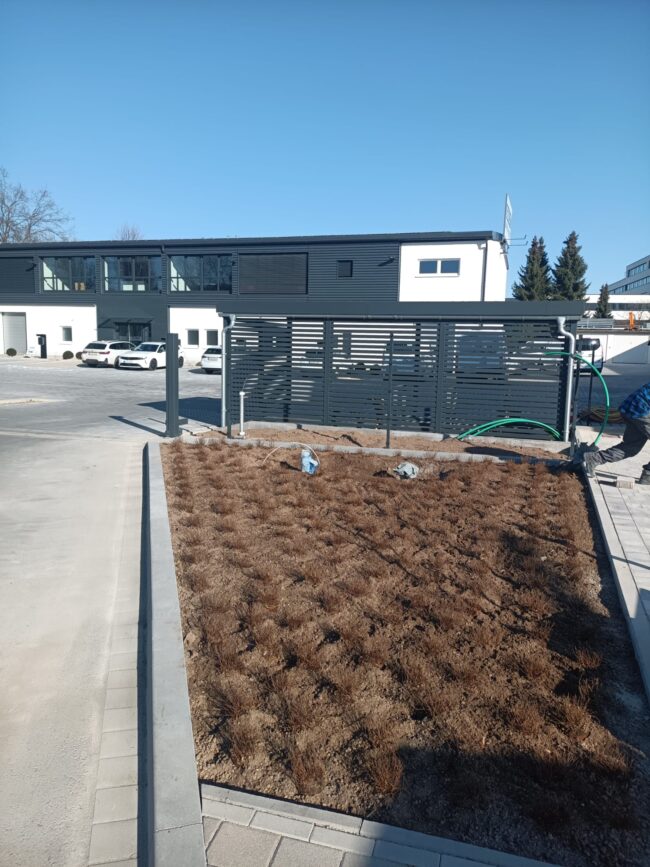 Elektro-Gerlitz in Bayreuth Neu angelegtes Firmengelände mit gepflasterter Zufahrt und einem großen Beet am Rand. Das Beet ist frisch bepflanzt mit kleinen Sträuchern in gleichmäßigem Raster. Im Hintergrund stehen Firmengebäude, Autos und eine Sichtschutzwand.