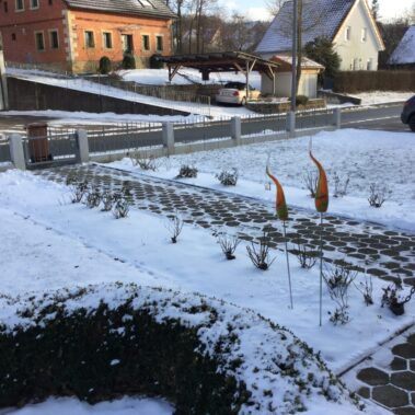 Vorgarten im Winter mit Schneedecke auf Rasen und Pflasterweg. Der Weg ist mit sechseckigen Steinen gepflastert.
