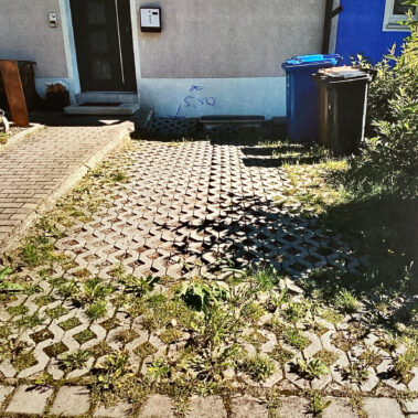 Eingangsbereich eines Hauses mit gepflasterter Einfahrt aus Rasengittersteinen, durch die Gras und Unkraut wachsen. Links führt ein kleiner Weg zur Haustür.